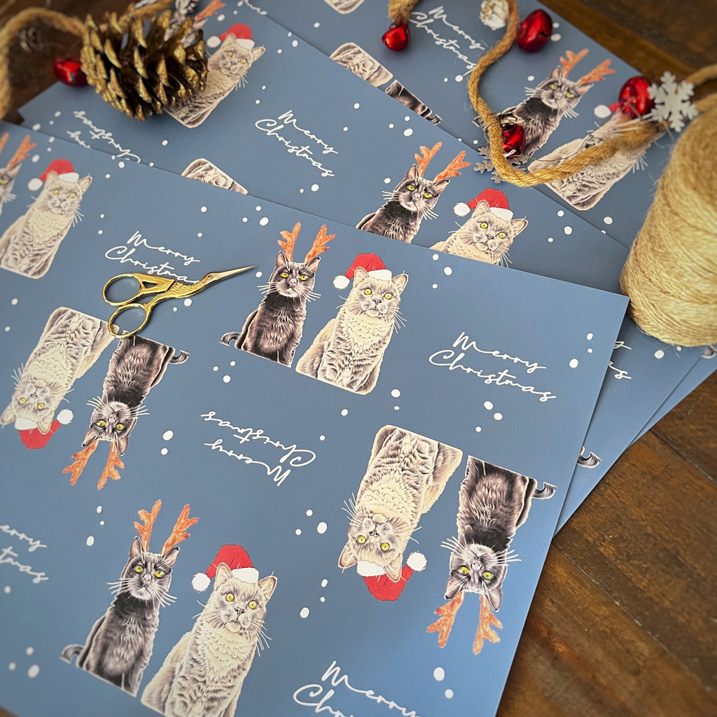 Blue Christmas-themed wrapping paper featuring illustrated cats wearing Santa hats and reindeer antlers, scattered on a wooden surface with holiday decorations including scissors, twine, a pinecone, and red ornaments.