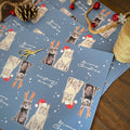 Blue Christmas-themed wrapping paper featuring illustrated cats wearing Santa hats and reindeer antlers, scattered on a wooden surface with holiday decorations including scissors, twine, a pinecone, and red ornaments.