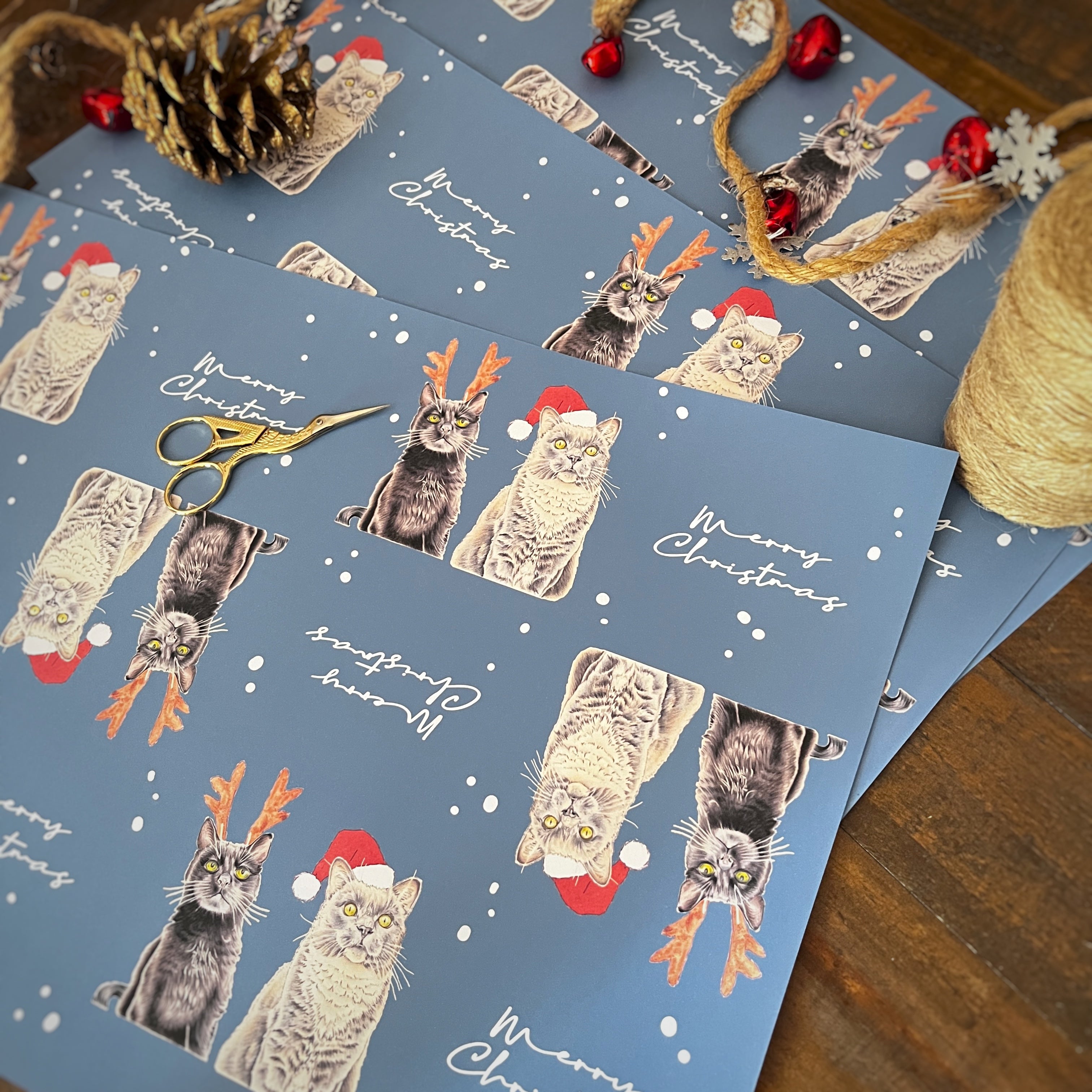 Blue Christmas-themed wrapping paper featuring illustrated cats wearing Santa hats and reindeer antlers, scattered on a wooden surface with holiday decorations including scissors, twine, a pinecone, and red ornaments.