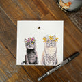A square greeting card featuring an  illustration black cat and grey cat wearing flower crowns, placed on a rustic wooden table with a black marker nearby.