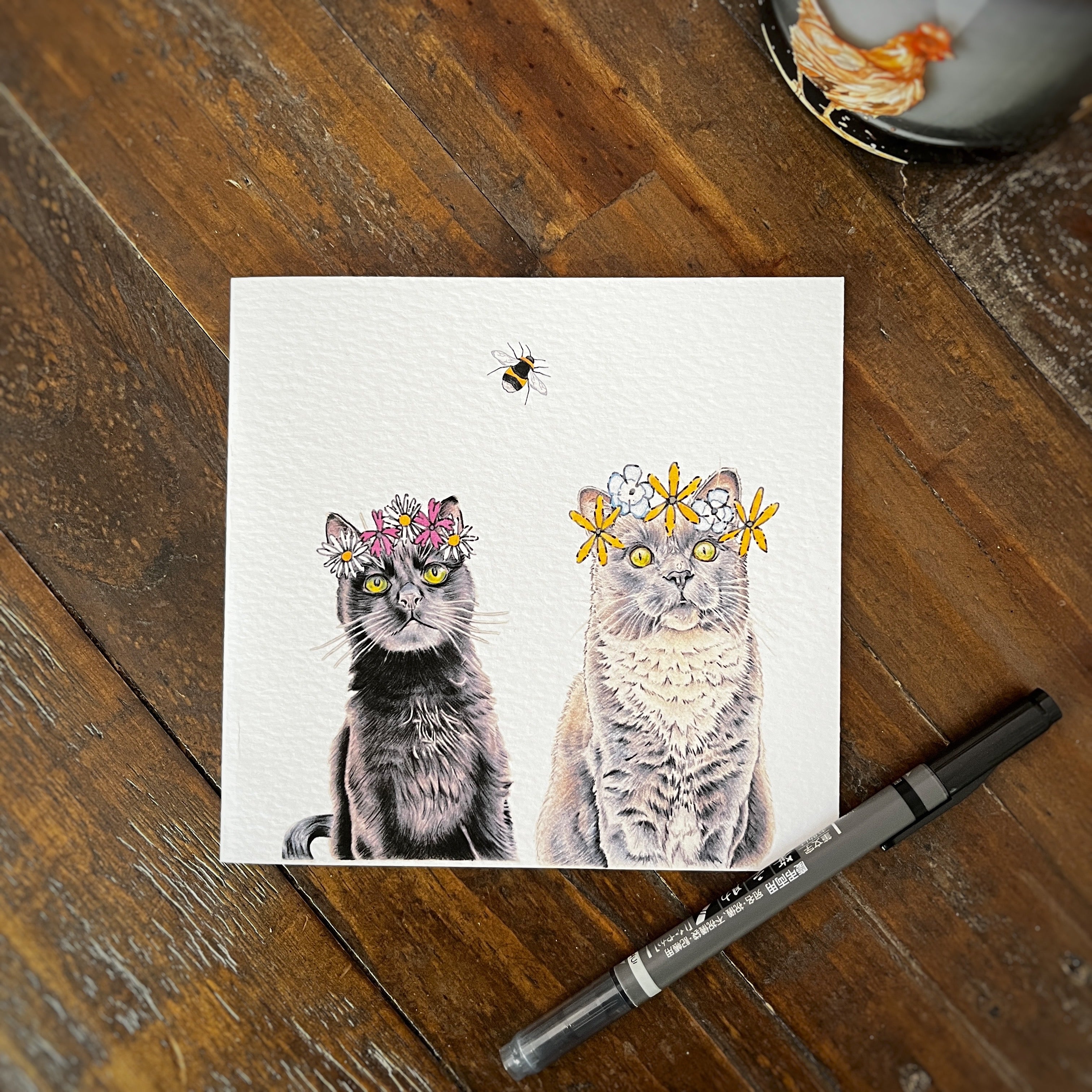 A square greeting card featuring an  illustration black cat and grey cat wearing flower crowns, placed on a rustic wooden table with a black marker nearby.