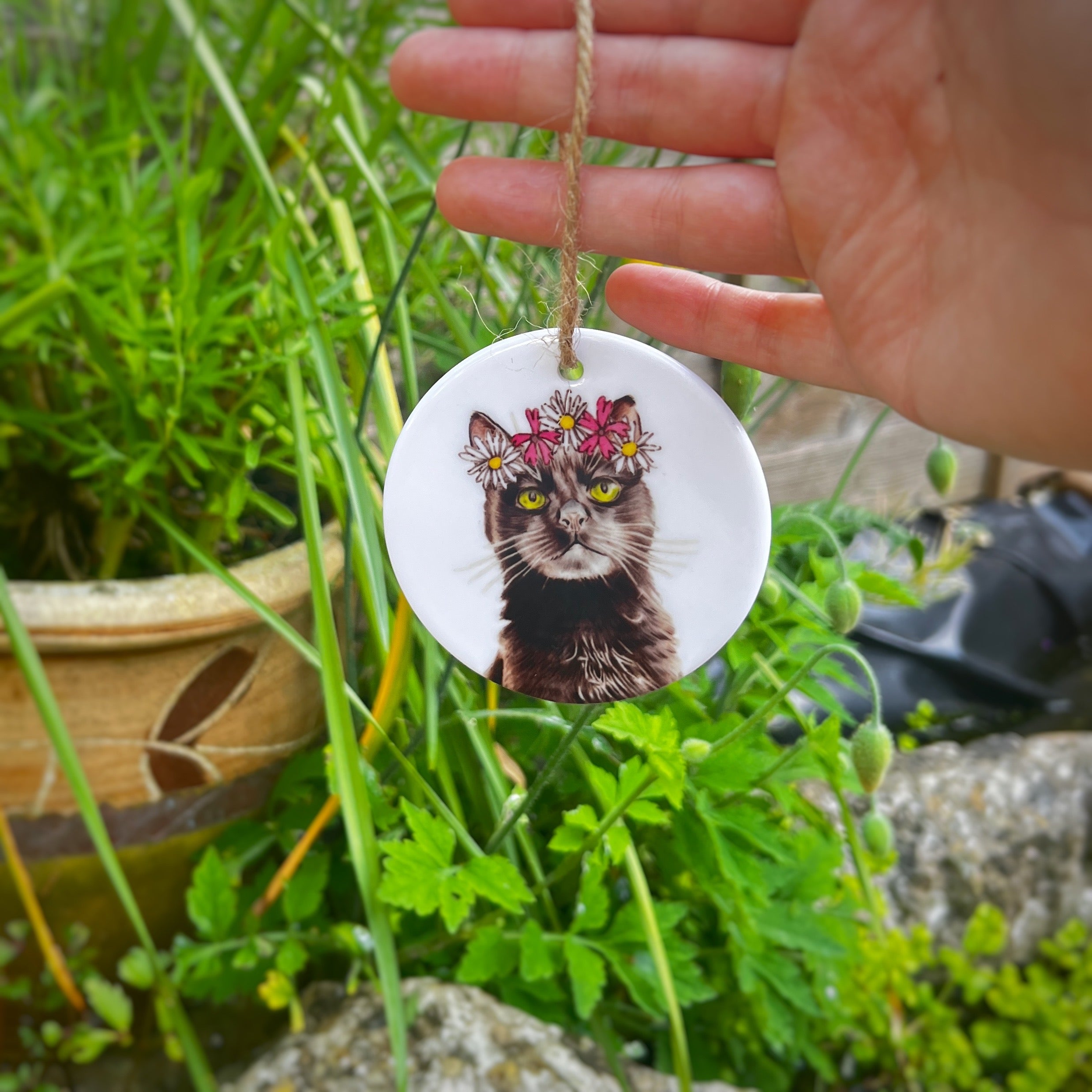 A hand holds a round white ceramic hanging ornament featuring a black cat portrait with a flower crown, outdoors in a garden setting with green plants and rocks in the background.