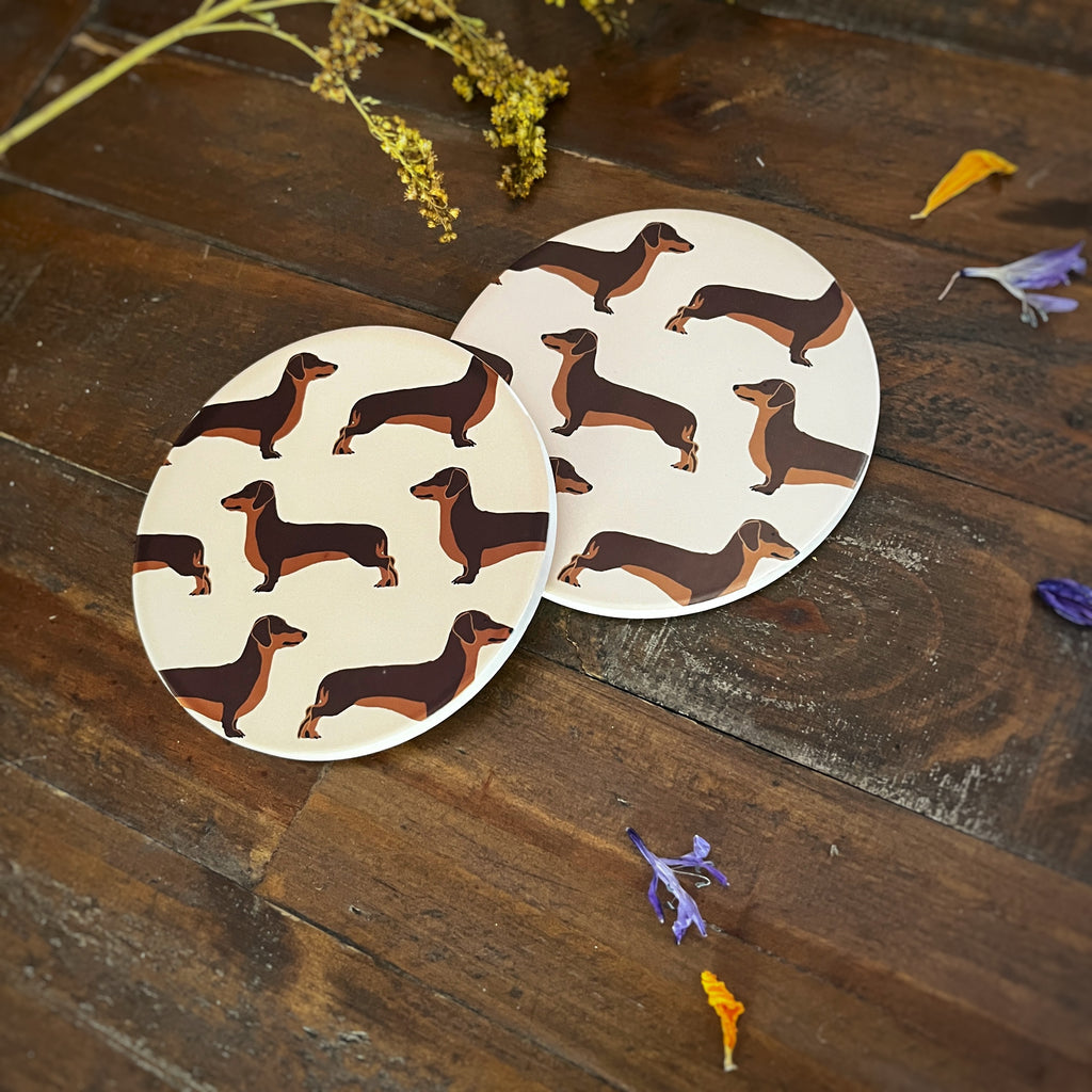 Two round coasters with a brown dachshund illustration pattern, displayed on a rustic dark wood surface with dried flowers.