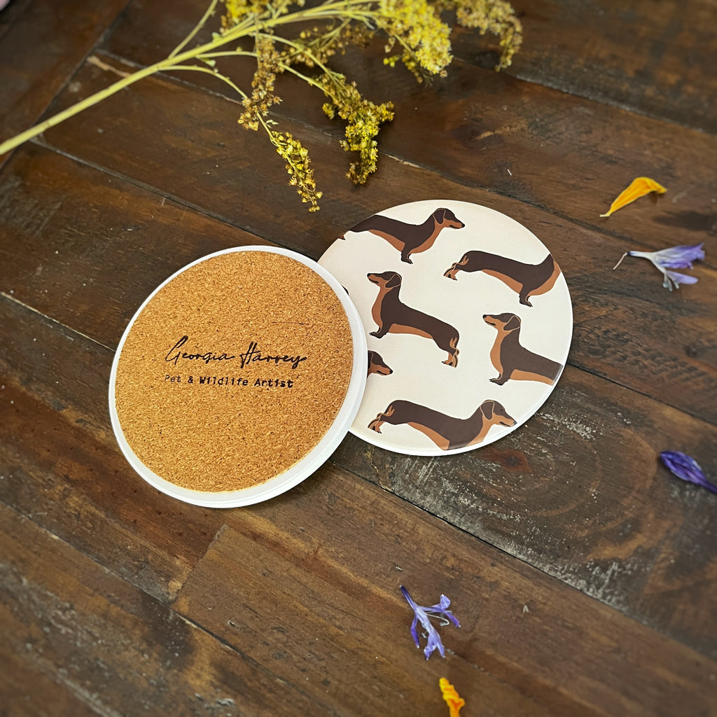 Two round brown sausage dog illustration pattern coasters on a dark wooden surface. One coaster shows a brown cork backing with a stamped logo 'Georgia Harvey Pet & Wildlife Artist'