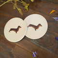 Two round coasters with a cream background, each printed with a brown dachshund illustration, displayed on a rustic dark wood surface with dried flowers.
