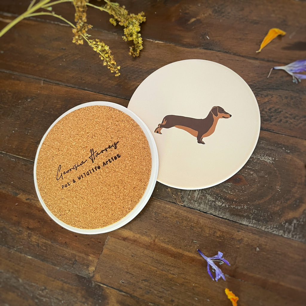 Two round coasters with a brown sausage dog illustration. One coaster shows a brown cork backing with the logo 'Georgia Harvey Pet & Wildlife Artist' stamped on it.