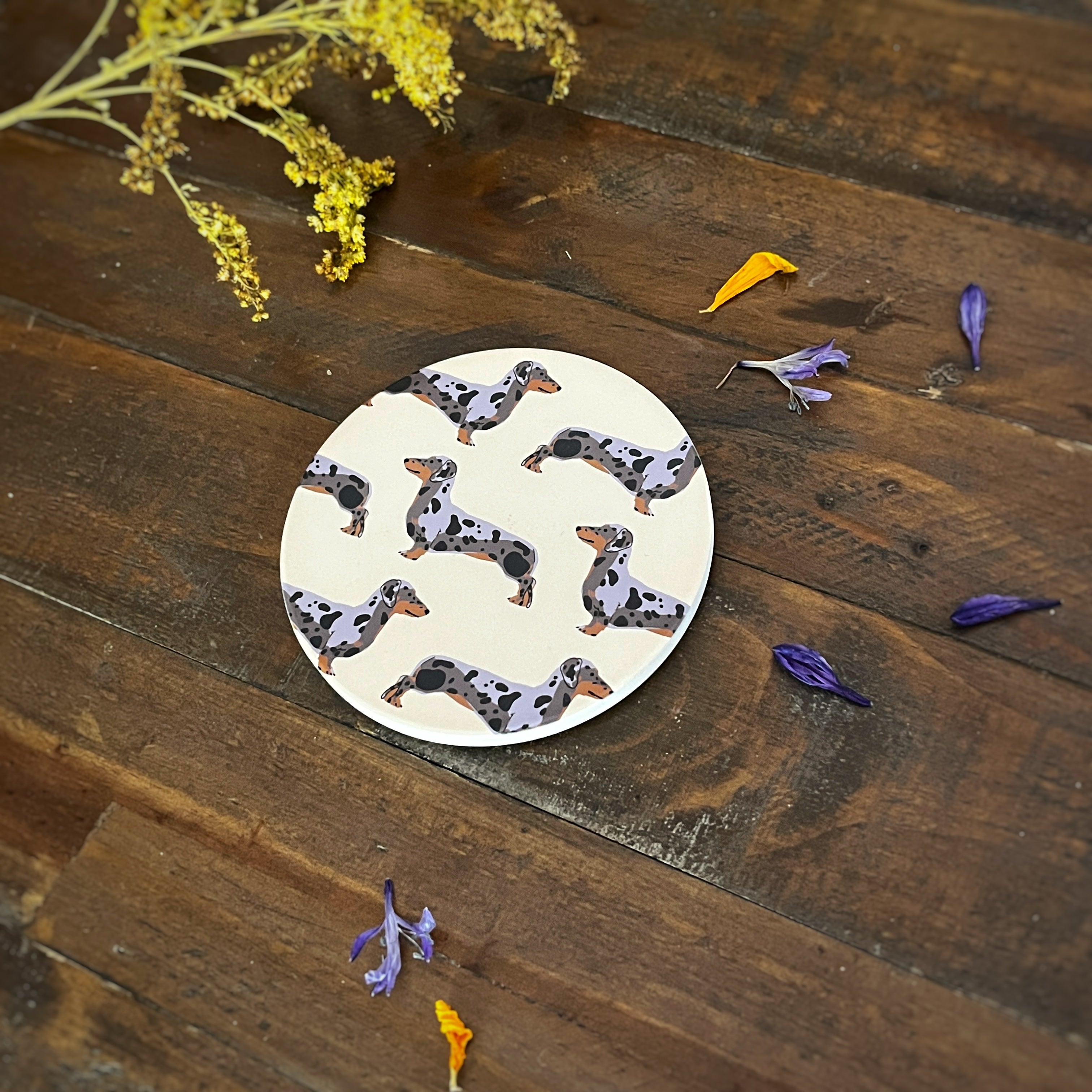 A round coaster featuring a repeating pattern of dapple dachshund dogs on a cream background, styled on a rustic dark wood surface with scattered purple and yellow flower petals.