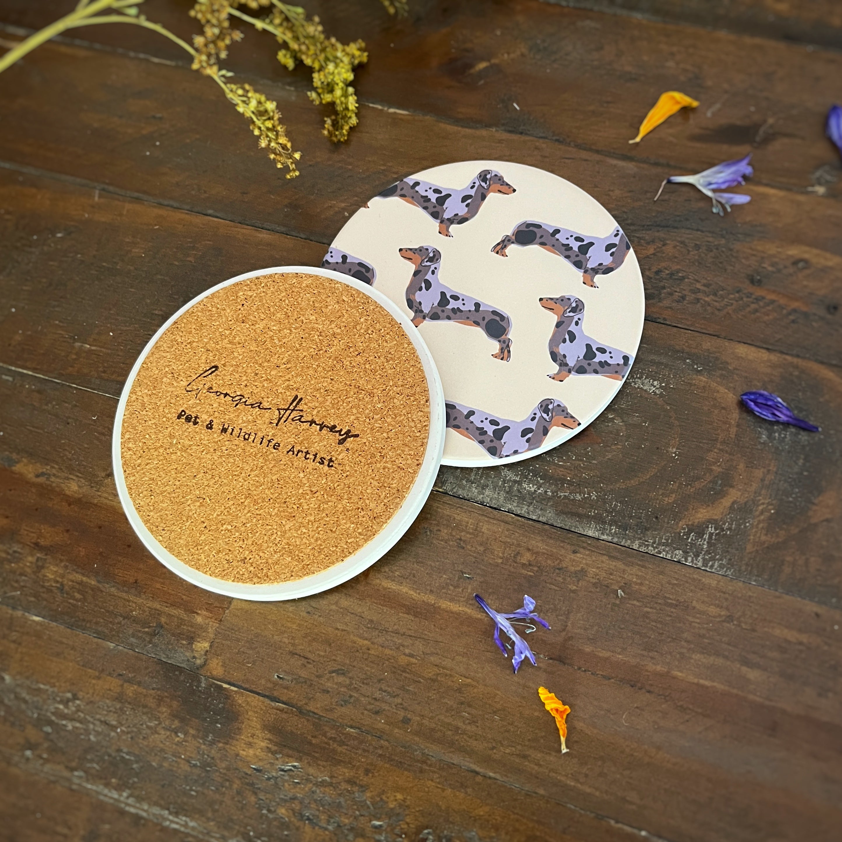 Two round coasters with dapple sausage dog illustration designs on a dark wooden tabletop. One coaster is showing a cork backing with a stamped logo 'Georgia Harvey Pet & Wildlife Artist'.
