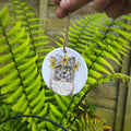 A round ceramic cat ornament with a printed illustration of a gray tabby cat wearing yellow and baby blue flowers, hanging from a twine string held by a hand in front of green fern leaves.