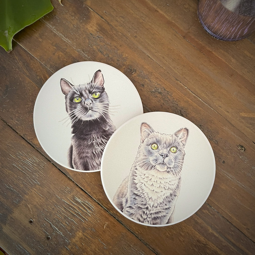 Two round white coasters featuring detailed illustrations of a black cat and gray cat with green eyes, displayed on a rustic wooden tabletop.