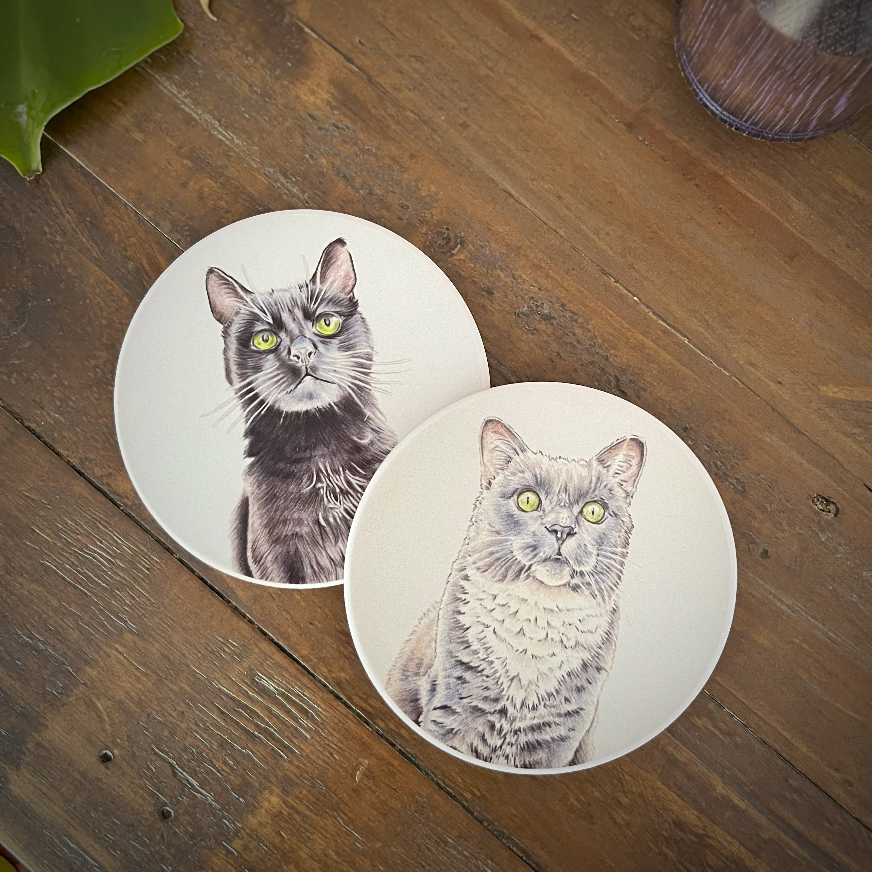 Two round white coasters featuring detailed illustrations of a black cat and gray cat with green eyes, displayed on a rustic wooden tabletop.