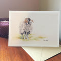 An A5 greeting card featuring a swaledale ram with curled horns, shown standing on a wooden surface with an envelope behind it and a purple quilted box in the background.