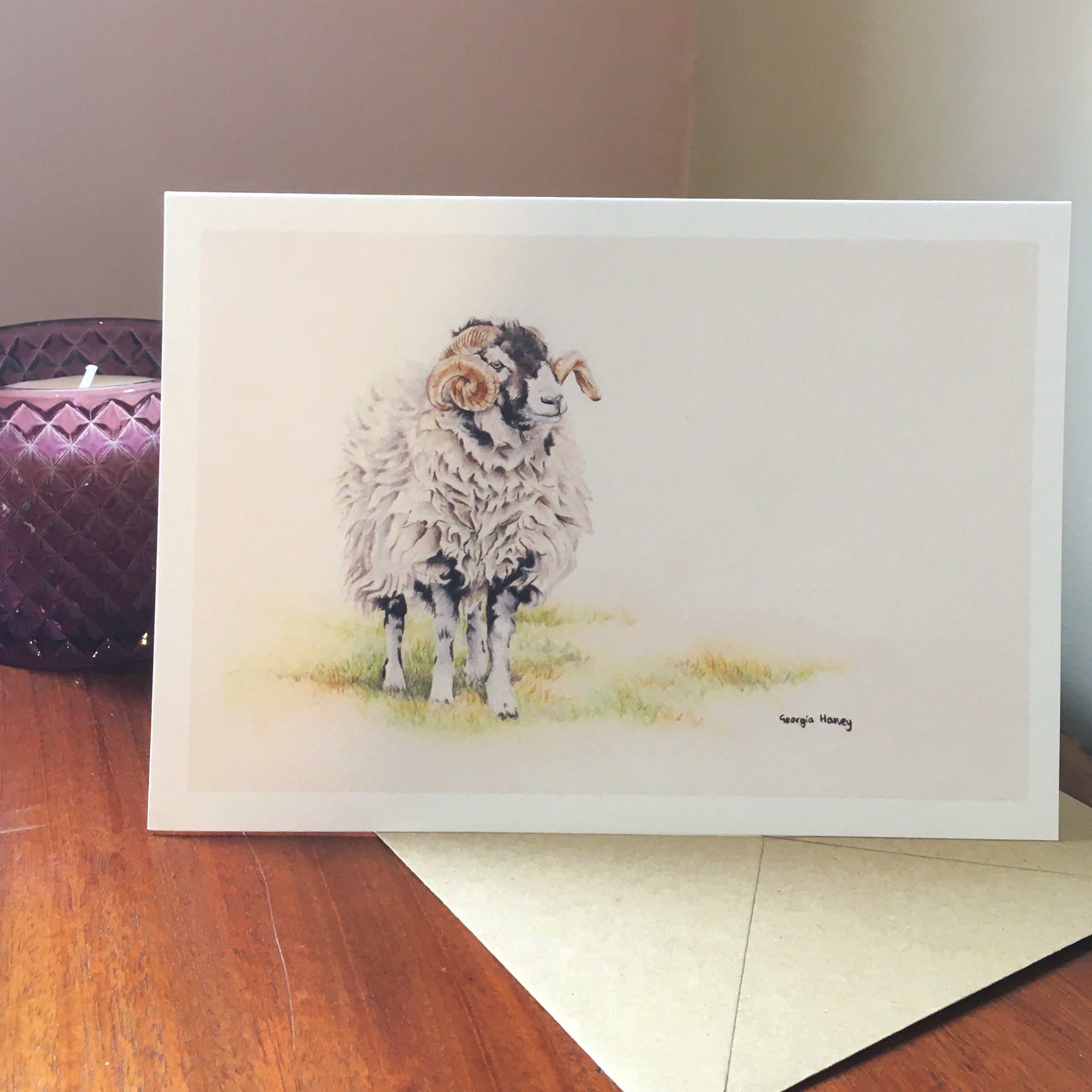 An A5 greeting card featuring a swaledale ram with curled horns, shown standing on a wooden surface with an envelope behind it and a purple quilted box in the background.