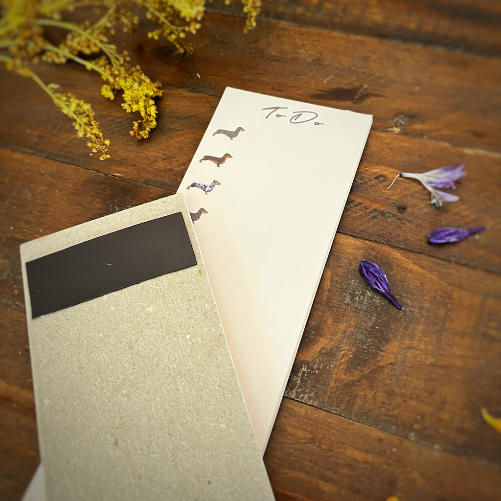 A cream-colored to-do list notepad with small sausage dog illustrations and a black magnetic cover, styled on a rustic wooden table with dried yellow flowers and purple petals.