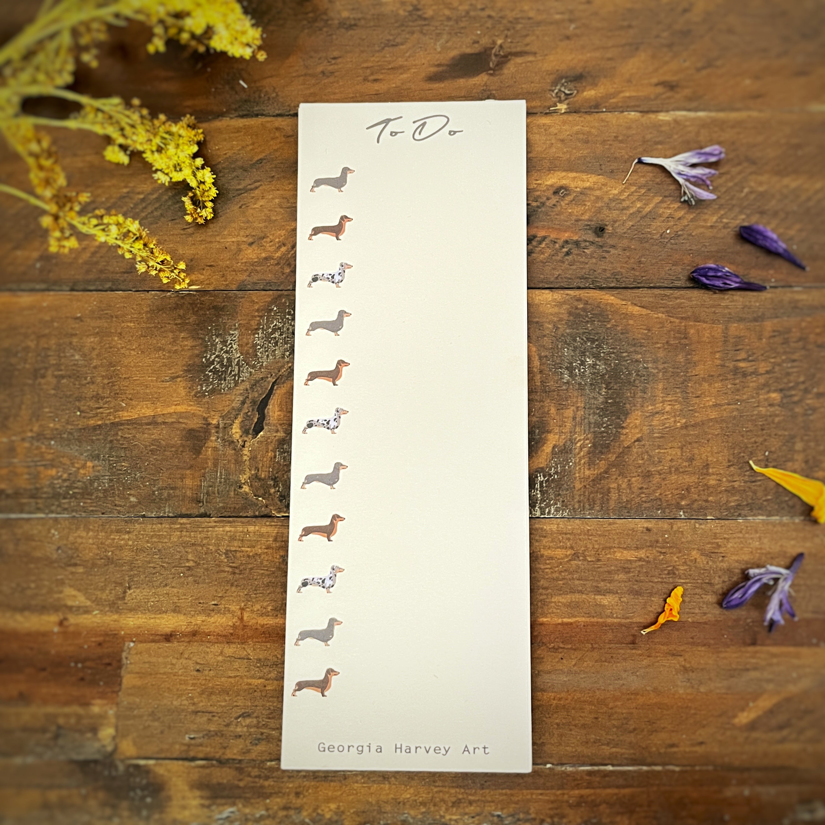 A vertical cream-colored list notepad titled 'To Do' with small illustrated dachshund figures and the name Georgia Harvey Art printed at the bottom, styled on a rustic wooden surface with scattered dried flowers.