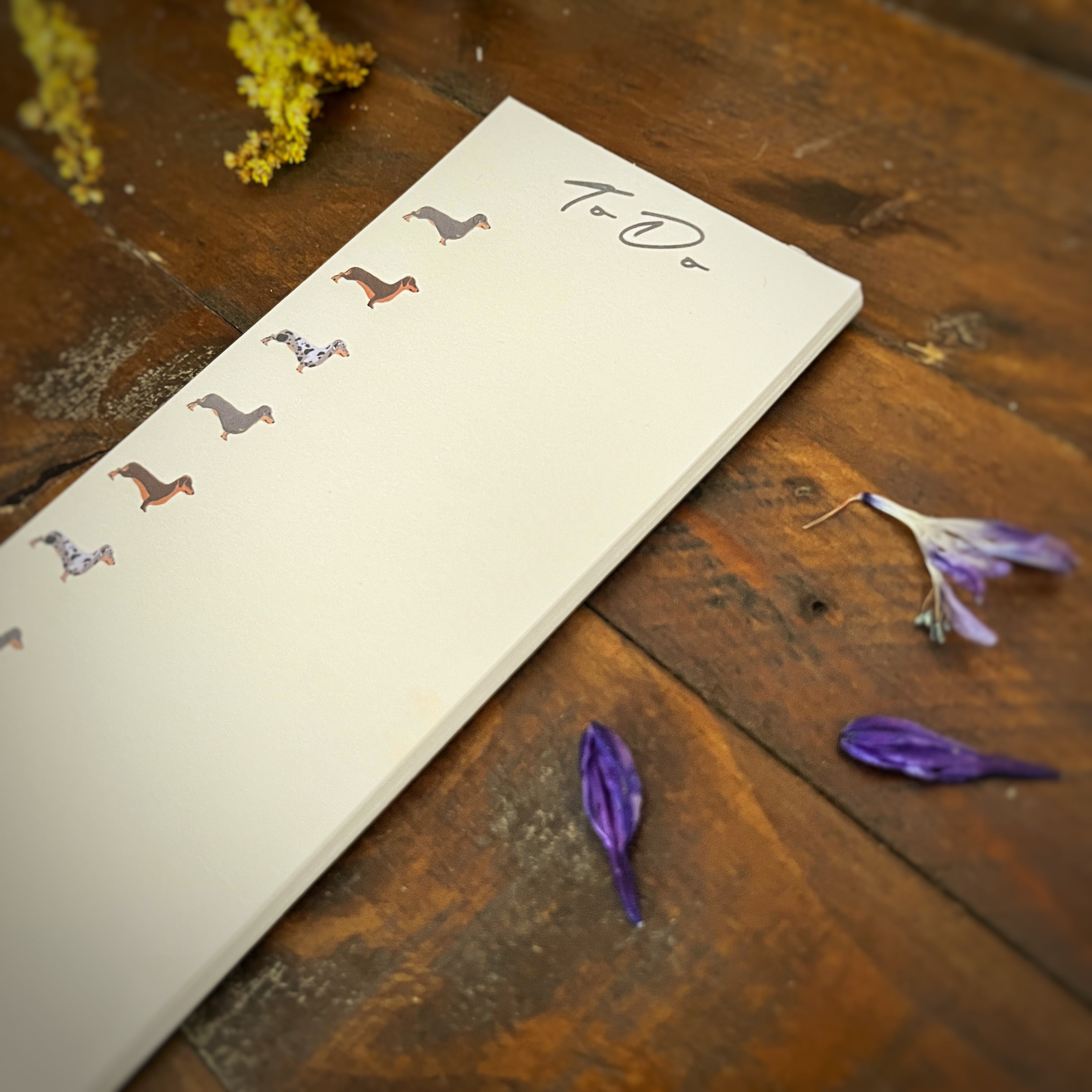A cream-colored list notepad with small illustrated dachshund illustrations along the left margin and the word 'To Do' written at the top, placed on a rustic wooden table with dried flowers scattered around.