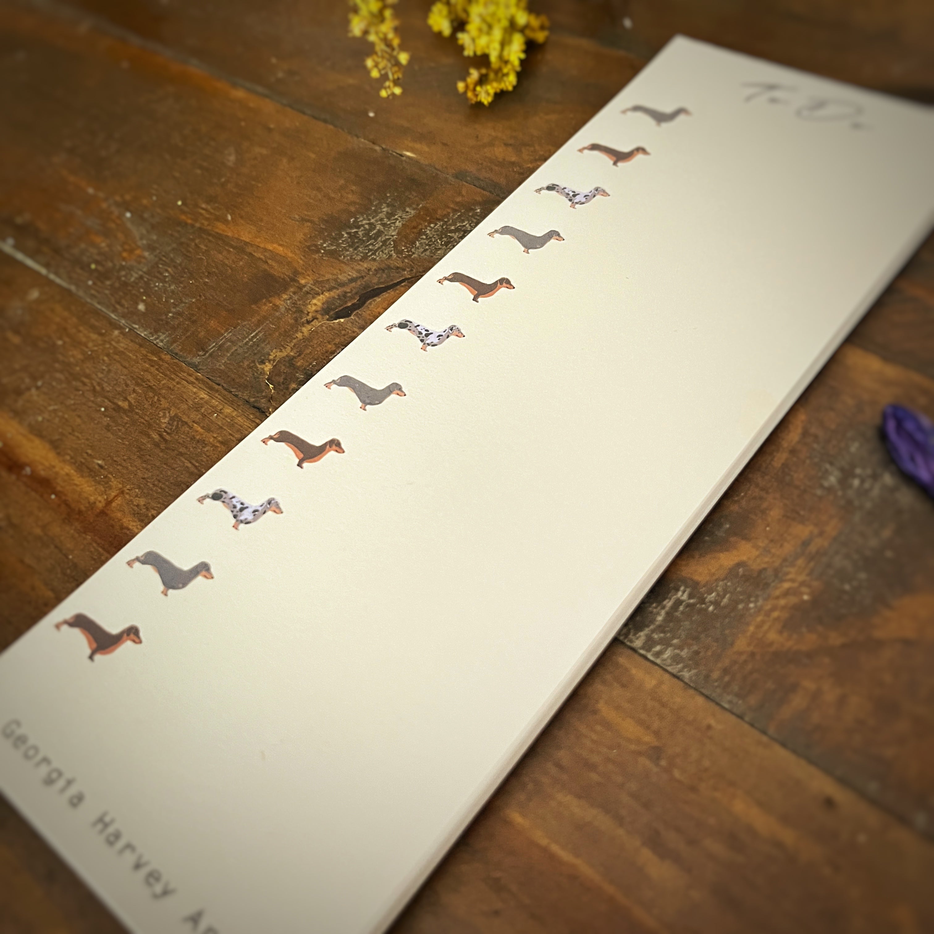 A vertical cream-colored list notepad lies diagonally on a rustic dark wood table. The notepad features a vertical row of small illustrated dachshund dogs in various colours, with the text 'To Do' at the top and 'Georgia Harvey Art' at the bottom.